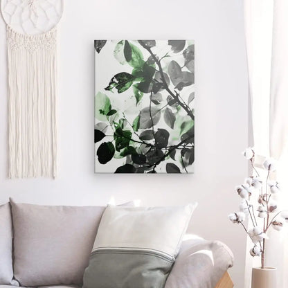 Une impression sur toile de feuilles vertes et noires, incarnant l'essence d'un Tableau Nature Feuilles Noir et Blanc Touche de Vert, est accrochée à un mur blanc au-dessus d'un canapé gris clair. A proximité, une tenture murale en macramé sublime l'Harmonie naturelle de la pièce aux côtés d'un vase en cotonnier.