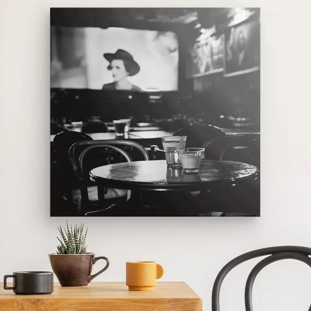 Une photo Tableau Cinéma Classique Scène Noir et Blanc capture un intérieur de café avec des chaises vides et une table ronde tenant des verres. En arrière-plan, un écran flou montre une personne portant un chapeau. Une table en bois ornée de tasses et d’une petite plante trône au premier plan, dégageant un air d’art décoratif.