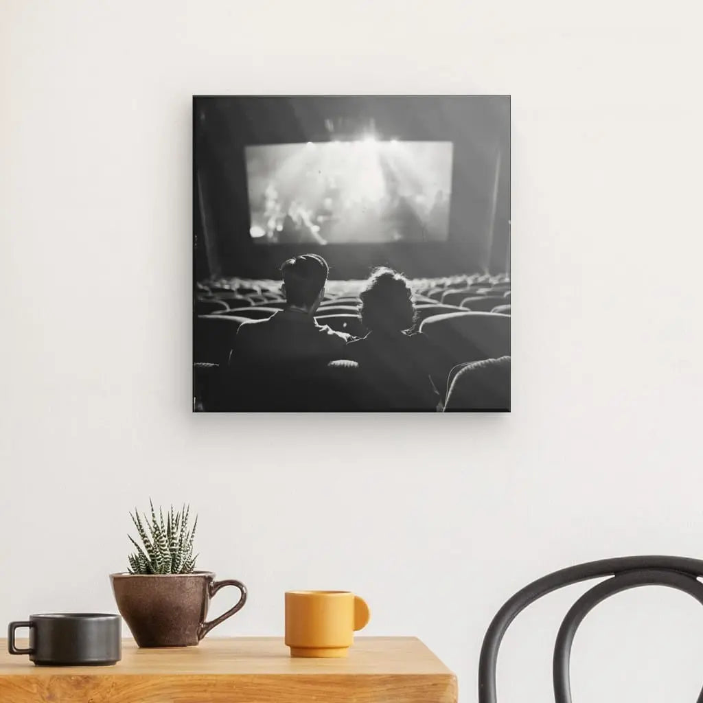 Une photo en noir et blanc sur un mur montre deux personnes assises dans une salle de cinéma, face à un écran lumineux, capturant un Tableau Cinéma Scène Ancienne Noir et Blanc. En dessous, il y a deux tasses, une petite plante et une chaise noire.