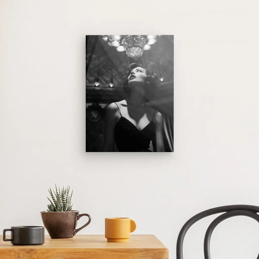 Une photo Noir et Blanc d'une femme vêtue d'une robe sombre, exsudant le glamour du Luxe des années 50 avec un lustre derrière elle. Ce Tableau Portrait Diva Femme 50's Luxe Noir et Blanc est accroché sur un mur blanc uni au-dessus d'une table en bois ornée d'une petite plante en pot et de deux tasses à café.