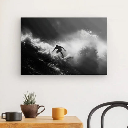 Une photographie en noir et blanc d'un surfeur intrépide surfant sur une vague est exposée sur un mur blanc au-dessus d'une table en bois avec une plante en pot, une tasse marron et une tasse jaune. Le Tableau Surf Océan Noir et Blanc capture l'essence de l'aventure. Une chaise noire est partiellement visible.