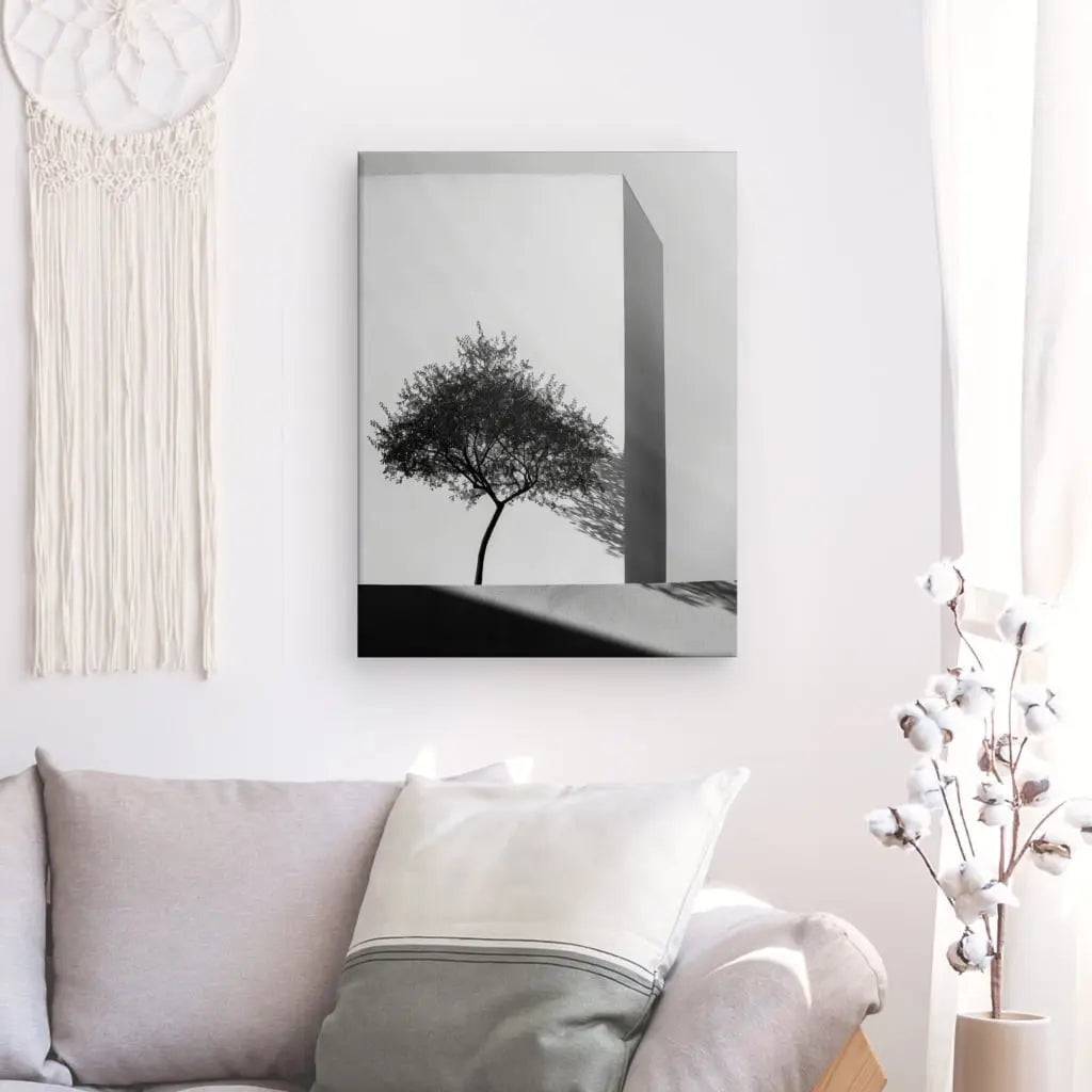 Photographie en noir et blanc d'un arbre projetant une ombre, encadrée et accrochée sur un mur blanc au-dessus d'un canapé gris clair avec un oreiller blanc. La décoration intérieure comprend un attrape-rêves et un vase de branches de coton, mettant en valeur le Tableau Architecture Naturelle Arbre Noir et Blanc comme une pièce maîtresse de l'art moderne.