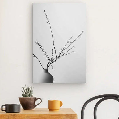 Un Tableau Nature Morte Pot Plante Branches Noir et Blanc photo d'un vase avec des branches sur un mur blanc au-dessus d'une table en bois avec une petite plante, une tasse noire et une tasse orange. Une chaise noire à dossier incurvé est partiellement visible à côté de la table, créant une scène artistique élégante.