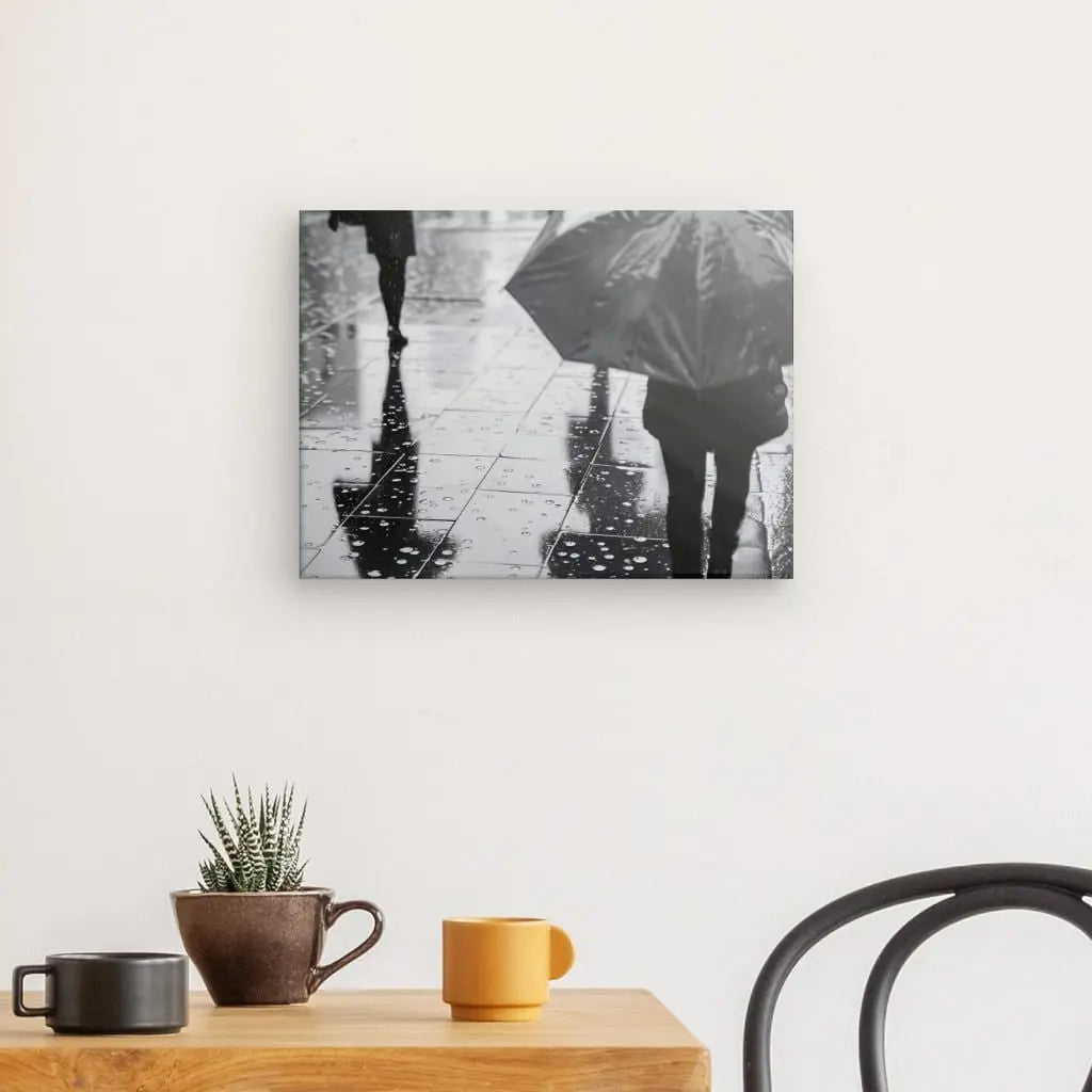 Le Tableau Pluie Parapluie Scène Urbaine Noir et Blanc représentant deux personnes marchant avec des parapluies sous la pluie est accroché sur un mur blanc au-dessus d'une petite table en bois avec deux tasses et une petite plante en pot.