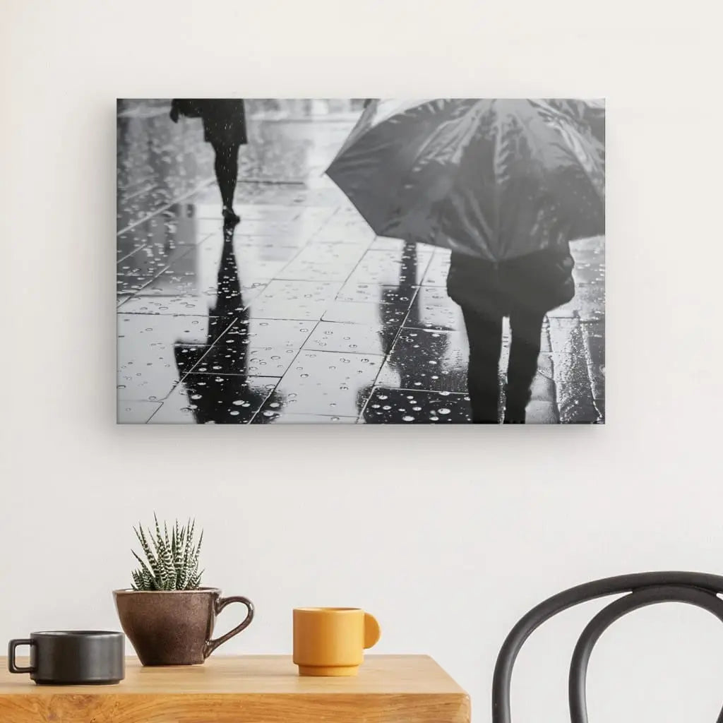 Un Tableau Pluie Parapluie Scene Urbaine Noir et Blanc capture une scène urbaine : des gens marchant dans une rue mouillée avec des parapluies, se reflétant dans les flaques d'eau. Cette photo saisissante est exposée sur un mur blanc au-dessus d'une table en bois ornée d'une petite plante et de deux tasses, mettant en valeur sa toile de haute qualité.