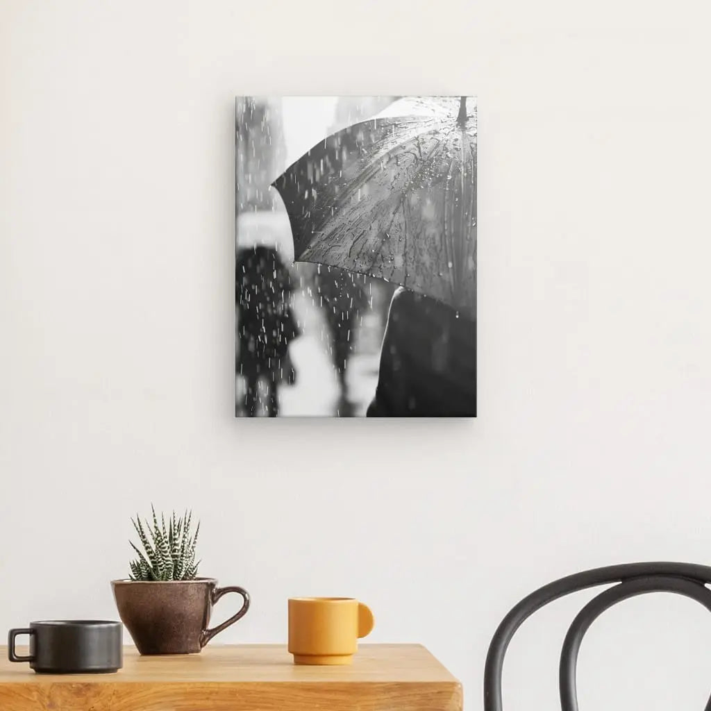 Un mur présente un Tableau Pluie Noir et Blanc, représentant une personne avec un parapluie sous la pluie. Ci-dessous, une table en bois avec deux tasses, une petite plante et une chaise sont disposées dans un décor minimaliste.