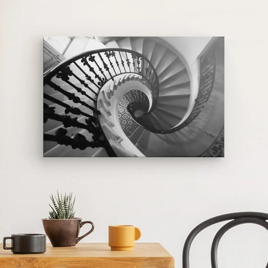 Photo en noir et blanc d'un escalier en colimaçon, encadrée comme une œuvre d'art et accrochée sur un mur blanc au-dessus d'une table en bois avec une petite plante en pot, une tasse marron et une tasse jaune. Une chaise noire est partiellement visible à droite. Ce Tableau Escalier Spirale Noir et Blanc ajoute de l'élégance à l'espace.