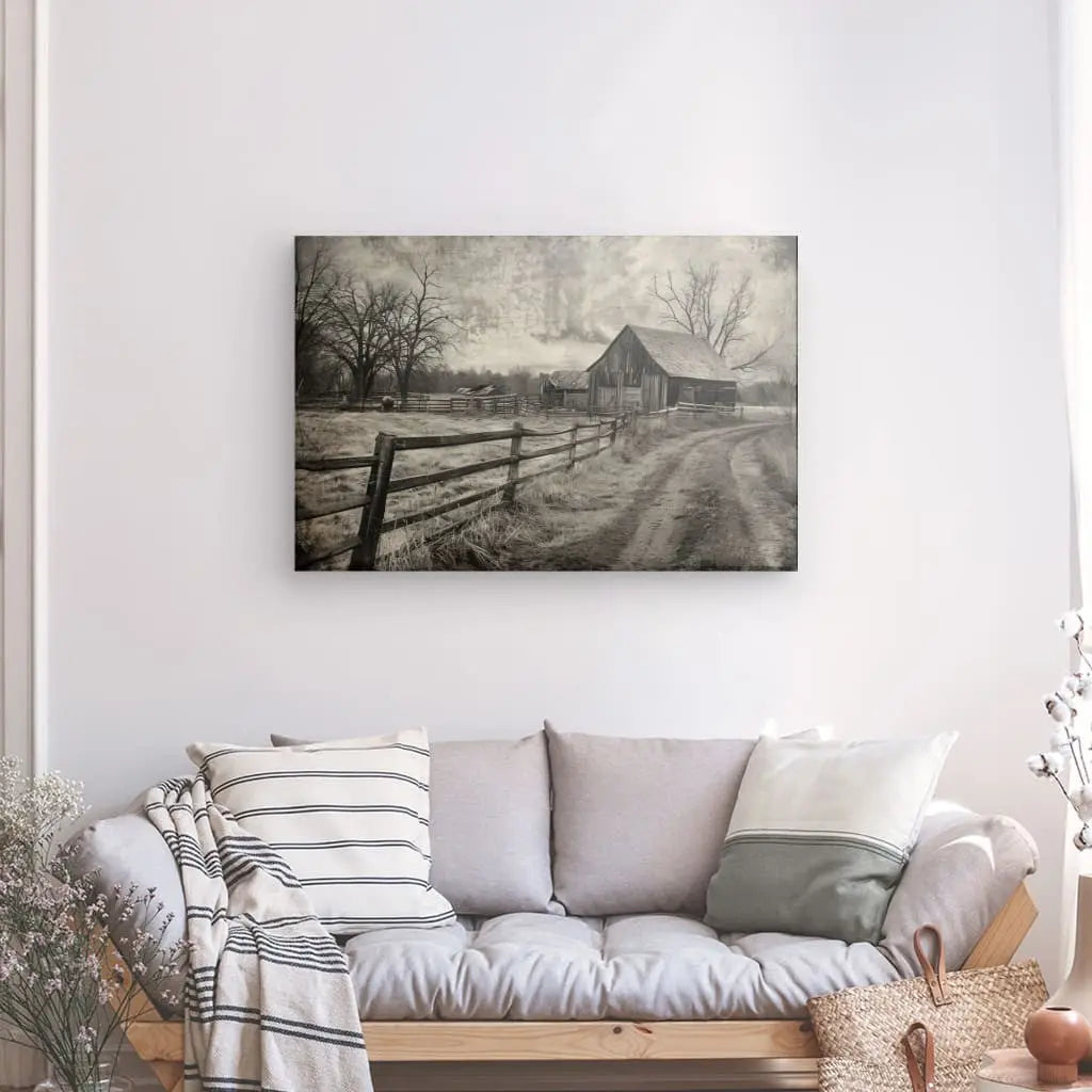 Une photographie en noir et blanc d’une grange rustique et d’un chemin de terre est accrochée à un mur blanc au-dessus d’un canapé gris clair avec des oreillers rayés et unis. Un panier en osier et des plantes sont posés sur le sol à côté du canapé, mettant en valeur le Tableau Paysage Campagne Ancien Noir et Blanc.