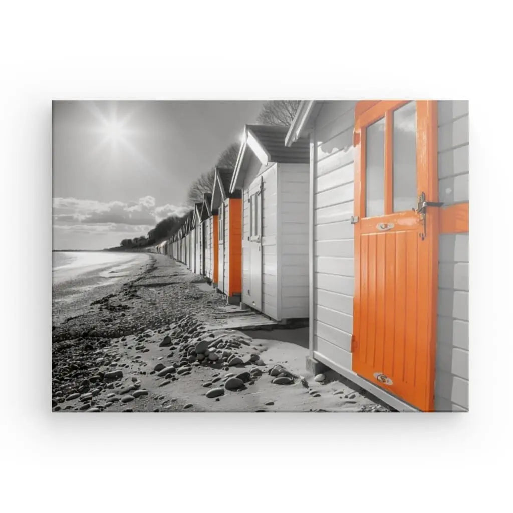 Une rangée de cabanes de bord de mer sur une plage de galets, représentées en noir et blanc, avec seules les portes orange. Le soleil brille dans un ciel partiellement nuageux, créant un captivant Tableau Cabane de Plage Noir Blanc Orange sur cette toile de haute qualité.