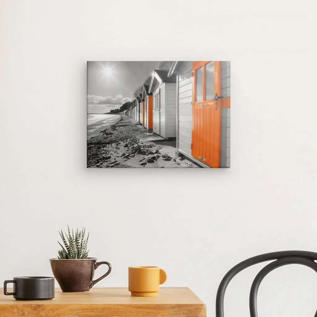 Un Tableau Cabane de Plage Noir Blanc Orange, avec les portes surlignées en orange, est accroché sur un mur blanc au-dessus d'une table en bois avec une plante en pot et deux tasses.