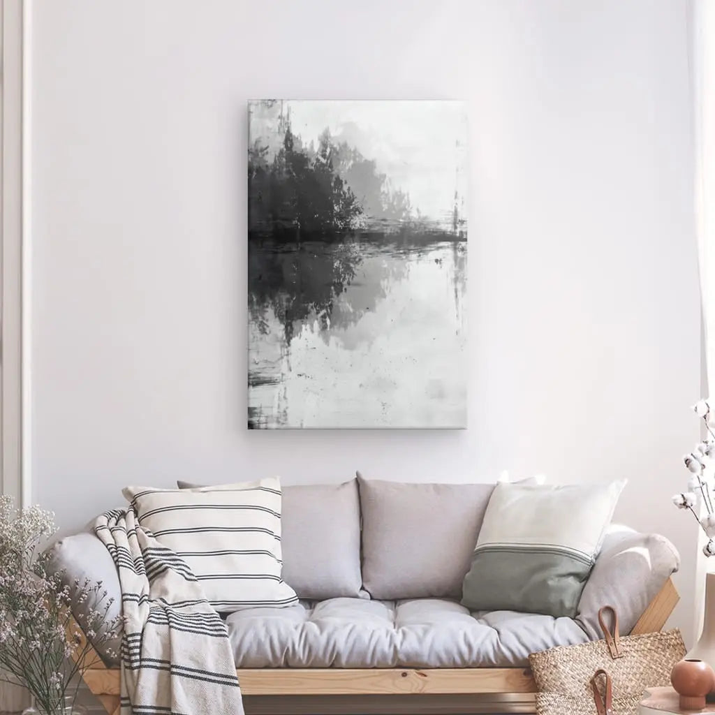 Un Tableau Abstrait Ombres Peinture Noir et Blanc d'arbres reflétés dans l'eau est suspendu sur un mur blanc au-dessus d'un canapé gris clair avec des coussins rayés et de couleur unie, à côté d'un panier et d'une plante en pot.