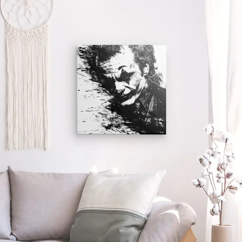 Une peinture monochrome représentant un homme avec du maquillage, rappelant le Joker de Batman, est accrochée sur un mur blanc au-dessus d'un canapé gris avec des coussins blancs et gris. Une décoration tissée et un vase avec des branches ajoutent à la décoration intérieure élégante de cet art Tableau Joker Batman Portrait Noir et Blanc.