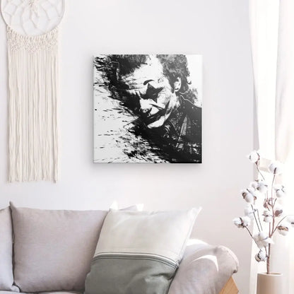 Une peinture monochrome représentant un homme avec du maquillage, rappelant le Joker de Batman, est accrochée sur un mur blanc au-dessus d'un canapé gris avec des coussins blancs et gris. Une décoration tissée et un vase avec des branches ajoutent à la décoration intérieure élégante de cet art Tableau Joker Batman Portrait Noir et Blanc.