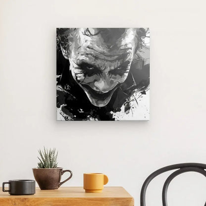 La peinture abstraite en noir et blanc représentant une personne avec un sourire sinistre ressemble au Tableau Peinture Joker Marvel Sourire Démoniaque Noir et Blanc sur un mur blanc au-dessus d'une table en bois avec une plante en pot, une tasse marron et une tasse jaune. Une partie d'une chaise noire est visible, soulignant l'allure étrange de cette œuvre d'art.