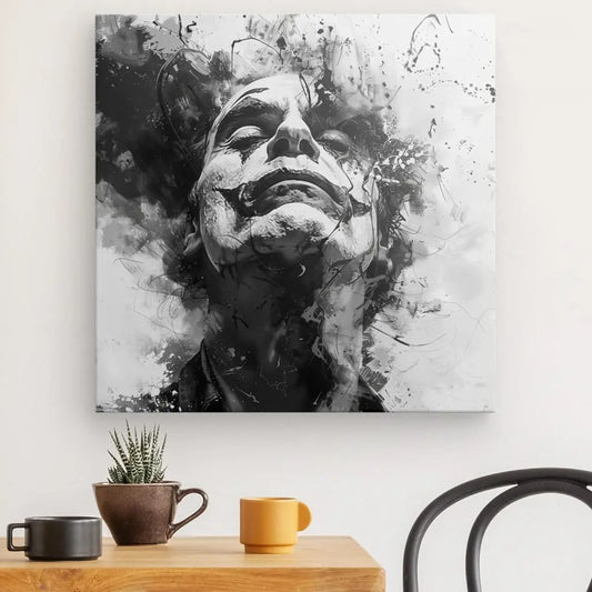 Tableau noir et blanc Joker Art Abstrait Noir et Blanc représentant une personne regardant vers le haut avec un fond abstrait et enfumé, accroché sur un mur blanc au-dessus d'une table en bois avec une plante en pot, une tasse marron et une tasse jaune.