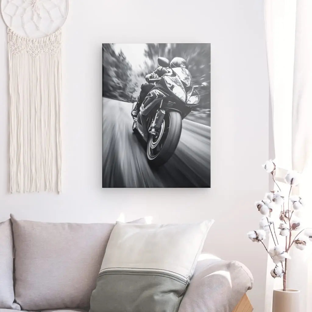 Photographie en noir et blanc d'une personne sur une moto de sport, montée sur un mur blanc au-dessus d'un canapé gris avec un coussin blanc. La décoration comprend une suspension murale en macramé blanc et un vase blanc avec des branches.