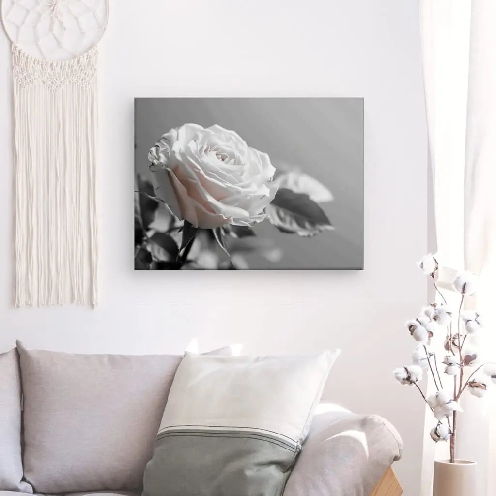 Un salon avec un canapé gris clair orné d'un oreiller blanc et gris. Un Tableau Rose Fleur Délicate Noir et Blanc sur toile tendue à la main est accroché au mur, à côté d'une tenture en macramé et d'un vase de branches de coton.