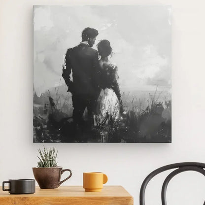 Un Tableau Couple Nature Peinture Noir et Blanc représentant un couple debout l'un près de l'autre, vu de dos. Cette décoration sophistiquée est suspendue au dessus d'une table en bois avec deux mugs, une petite plante et une chaise à proximité.