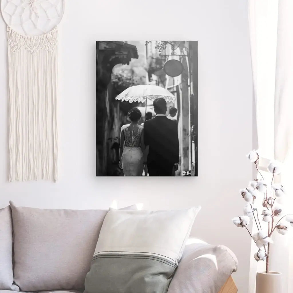 Une photo en noir et blanc de deux personnes sous un parapluie, vues de dos, est accrochée sur un mur blanc au-dessus d'un canapé moderne avec des coussins gris clair. Cette œuvre d'art romantique est complétée par un décor de cotonnier à côté du canapé.