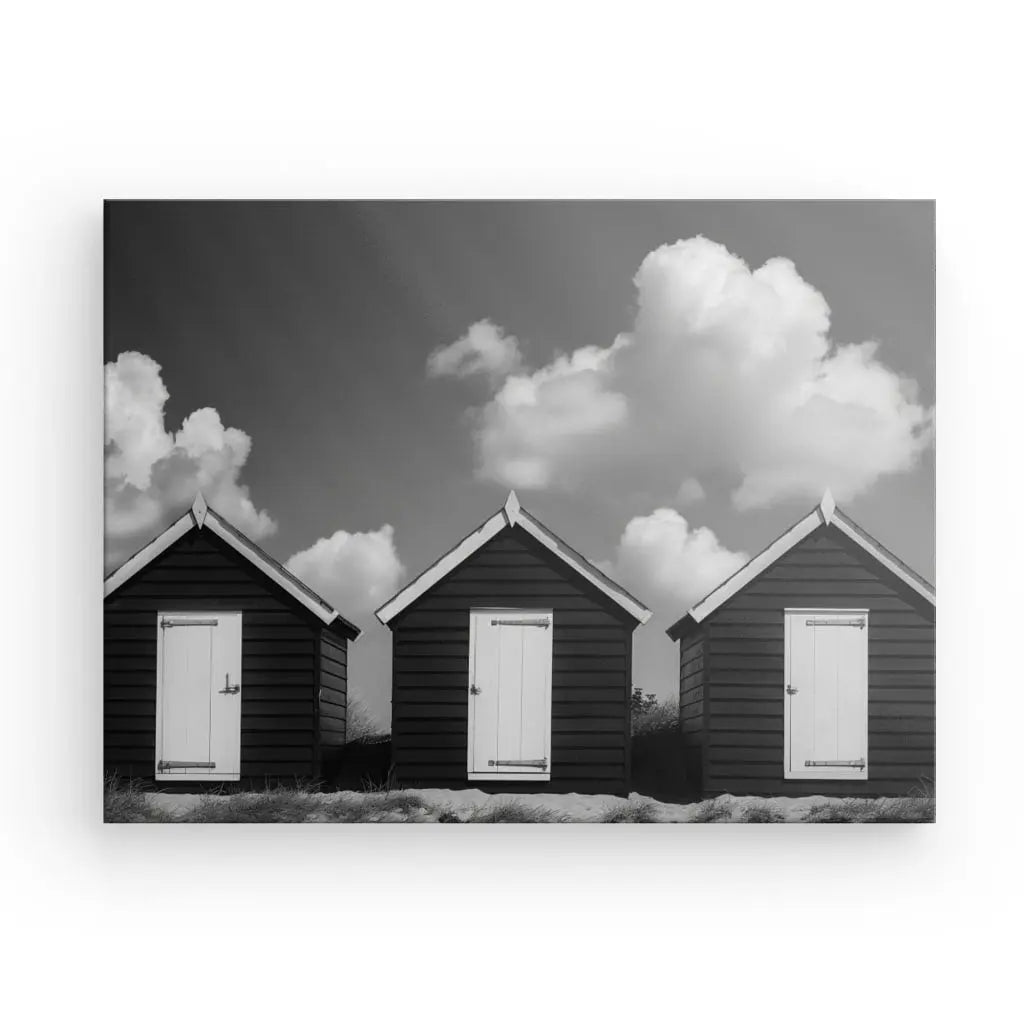 Trois petites cabanes en bois sombre avec des portes et des boiseries blanches s'alignent sous un ciel nuageux, créant un pittoresque Tableau Cabanes de Plage Noir et Blanc. Cette scène dégage un sentiment de décoration élégante, incarnant l'art côtier intemporel.