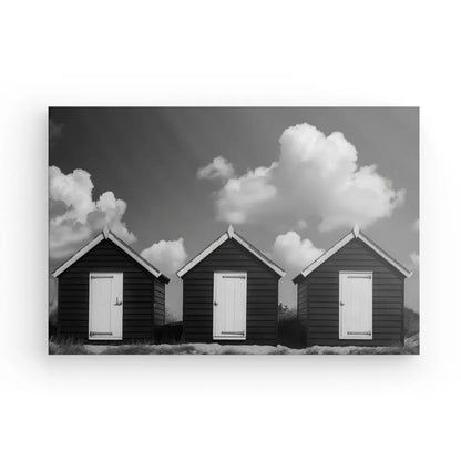 Trois petites cabanes de plage en bois sombre avec des portes blanches s'alignent sur un fond de nuages épars sur une photographie en noir et blanc. Cette œuvre d'art saisissante, Tableau Cabanes de Plage Noir et Blanc, ajoute une touche de décoration élégante et constitue un côtier d'art idéal.