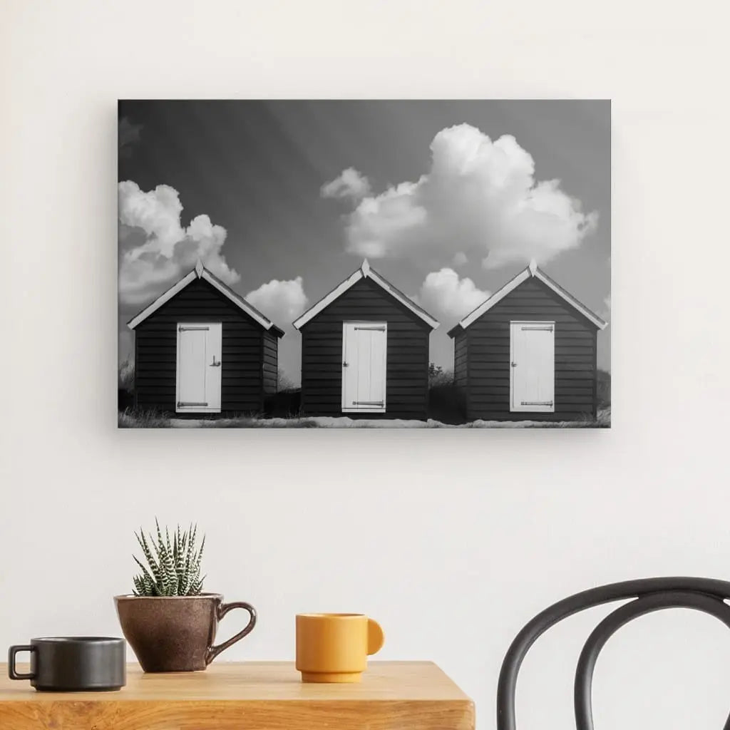 Une photo en noir et blanc, Tableau Cabanes de Plage Noir et Blanc, de trois petites cabanes de plage en bois accrochées à un mur blanc ajoute une touche de décoration élégante. Au premier plan, une table en bois avec une plante verte en pot, une tasse marron et une tasse orange complètent cette ambiance art côtier.