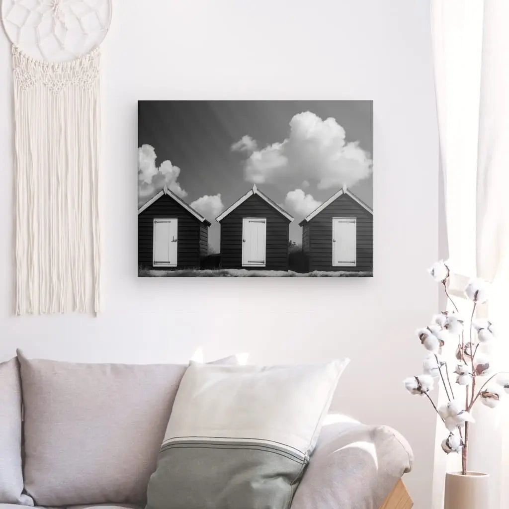 Photo en noir et blanc de trois petites maisons sur un mur au-dessus d'un canapé gris, avec une tenture murale en macramé et un vase avec des branches de coton à proximité, créant une ambiance de décoration élégante.