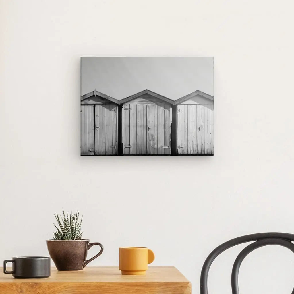 Un mur avec une photo en noir et blanc Tableau Cabanons Plage Sable Noir et Blanc de trois hangars en bois, au-dessus d'une table en bois qui contient une petite plante en pot et trois tasses dépareillées. Une chaise partiellement noire est visible sur le côté droit, ajoutant à l'ambiance photographique artistique décoration côtière.