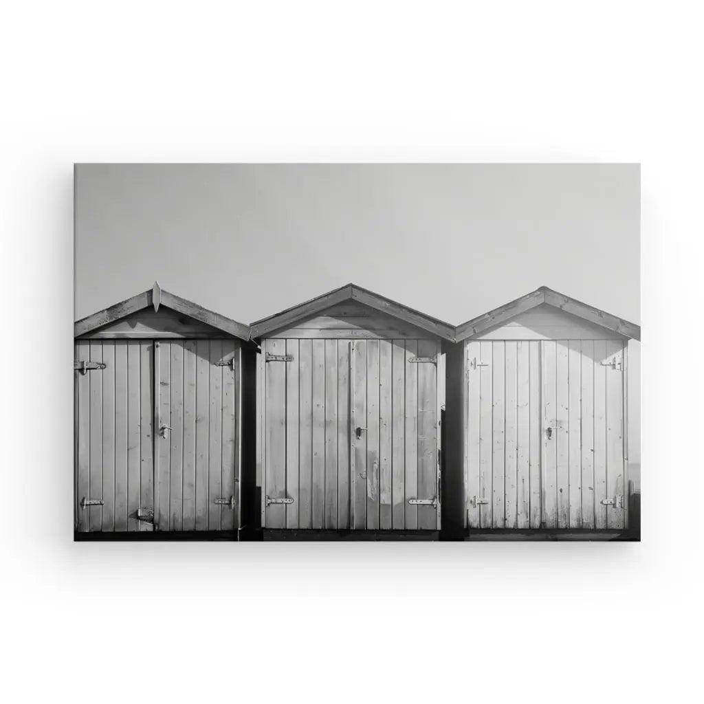 Trois hangars de stockage en bois en enfilade, chacun avec un toit en pente et une porte fermée. L'image en noir et blanc met en valeur la texture et les détails des planches de bois, créant une photographie artistique rappelant le Tableau Cabanons Plage Sable Noir et Blanc.