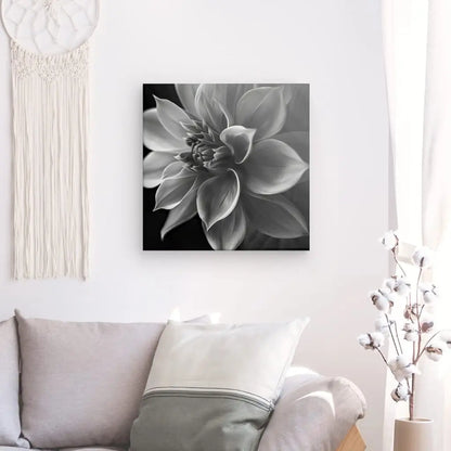 Une toile monochrome intitulée "Fleur Lotus Gros Plan Noir et Blanc" est accrochée à un mur blanc au-dessus d'un canapé beige orné de coussins de couleur claire. Un rideau blanc recouvre partiellement une fenêtre et une branche de coton complète le décor, rehaussant le décor zen.