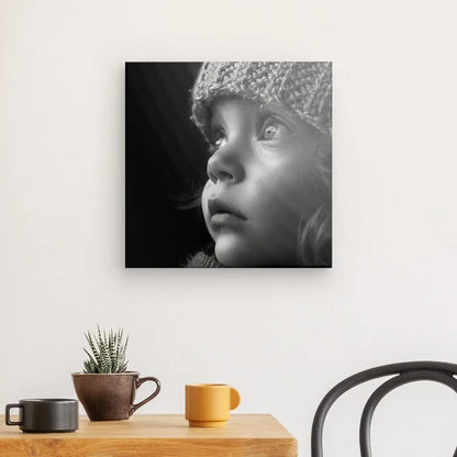 Un Tableau Portrait Enfant Bonnet Noir et Blanc est accroché sur un mur blanc au-dessus d'une table en bois. Sur la table se trouvent un petit cactus, une tasse marron et une tasse jaune.