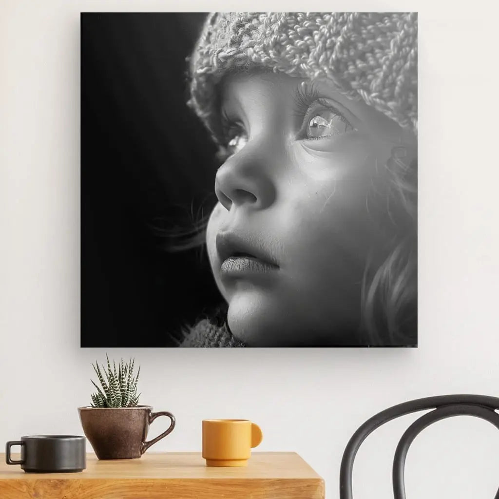 Photo en noir et blanc d'un visage d'enfant portant un chapeau d'hiver, affichée sous forme de Tableau Portrait Enfant Bonnet Noir et Blanc sur un mur blanc au-dessus d'un bureau en bois avec une plante en pot, une tasse marron et une tasse jaune. Une chaise noire est partiellement visible.