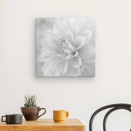 Un Tableau Fleur Blanche Pétales Noir et Blanc est accroché sur un mur d'un blanc immaculé. Ci-dessous, une petite table contient une tasse brune, une tasse jaune et une petite plante en pot. Une chaise noire est partiellement visible à droite, contribuant à la décoration minimaliste.