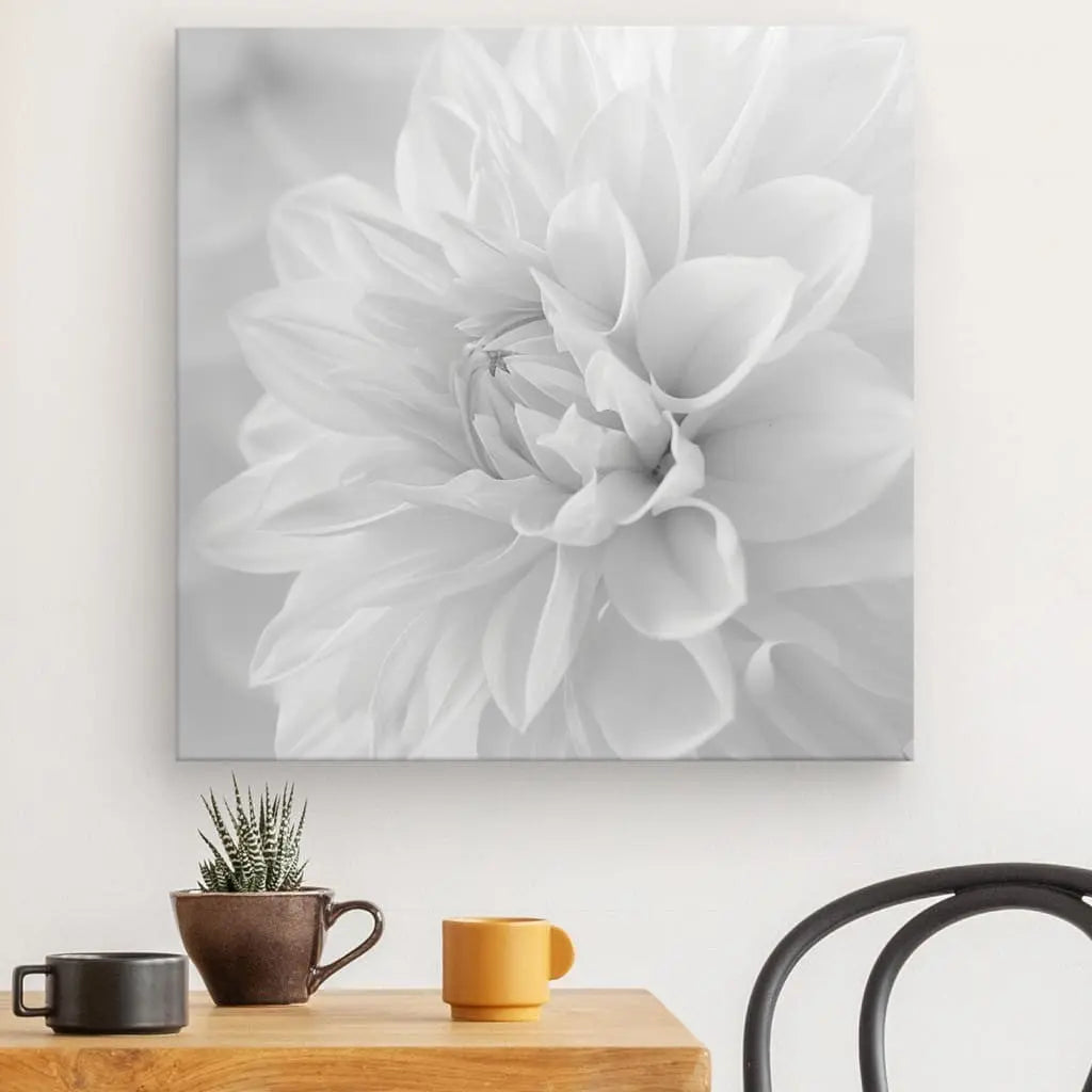 Un Tableau Fleur Blanche Pétales Noir et Blanc est accroché au mur au-dessus d'une table en bois. Le décor minimaliste comprend une petite plante en pot, une tasse noire et une tasse jaune. Une chaise noire est partiellement visible à côté de la table, renforçant la décoration minimaliste.