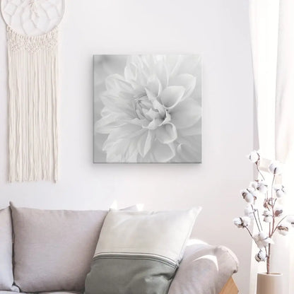 Un Tableau Fleur Blanche Pétales Noir et Blanc présentant une photo en niveaux de gris d'une fleur blanche est accroché sur un mur blanc au-dessus d'un canapé gris clair avec un coussin pâle. A gauche, une tenture murale en macramé, et à droite, un vase aux branches blanches ajoute à la décoration minimaliste.