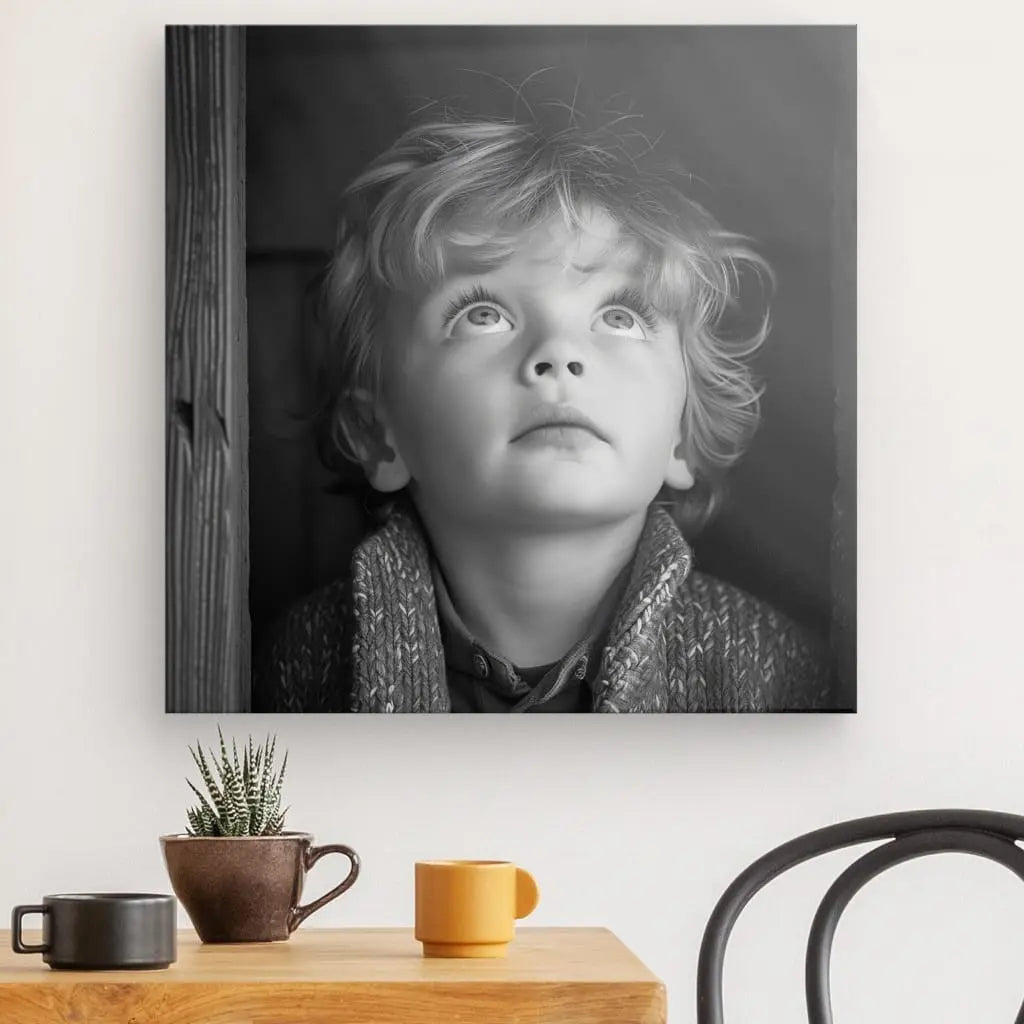 Un saisissant Tableau Jeune Garçon Tête Blonde Noir et Blanc capture un jeune garçon regardant vers le haut, encadré comme une photographie artistique sur le mur. En dessous, deux mugs et un petit cactus en pot reposent sur la table.