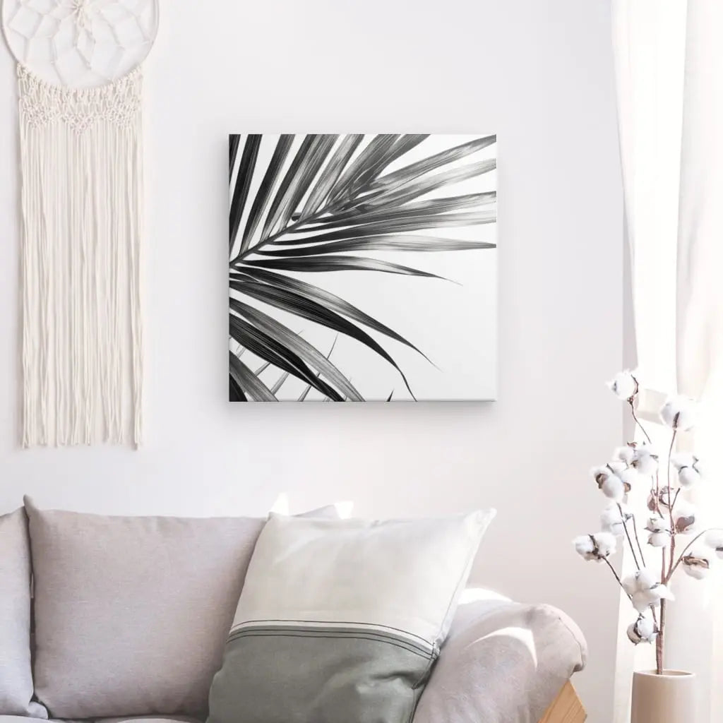 Un salon minimaliste comprend un canapé gris, des oreillers blancs et gris, une peinture de feuilles de palmier en noir et blanc sur le mur, un attrape-rêves et un vase blanc avec des branches de coton sur une table d'appoint. Le décor reflète l'élégance contemporaine avec des touches comme le Tableau Fougères Feuilles Minimalistes Noir et Blanc qui rehaussent l'esthétique.