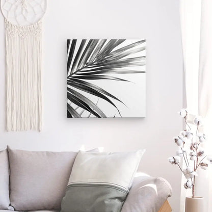 Un salon minimaliste comprend un canapé gris, des oreillers blancs et gris, une peinture de feuilles de palmier en noir et blanc sur le mur, un attrape-rêves et un vase blanc avec des branches de coton sur une table d'appoint. Le décor reflète l'élégance contemporaine avec des touches comme le Tableau Fougères Feuilles Minimalistes Noir et Blanc qui rehaussent l'esthétique.