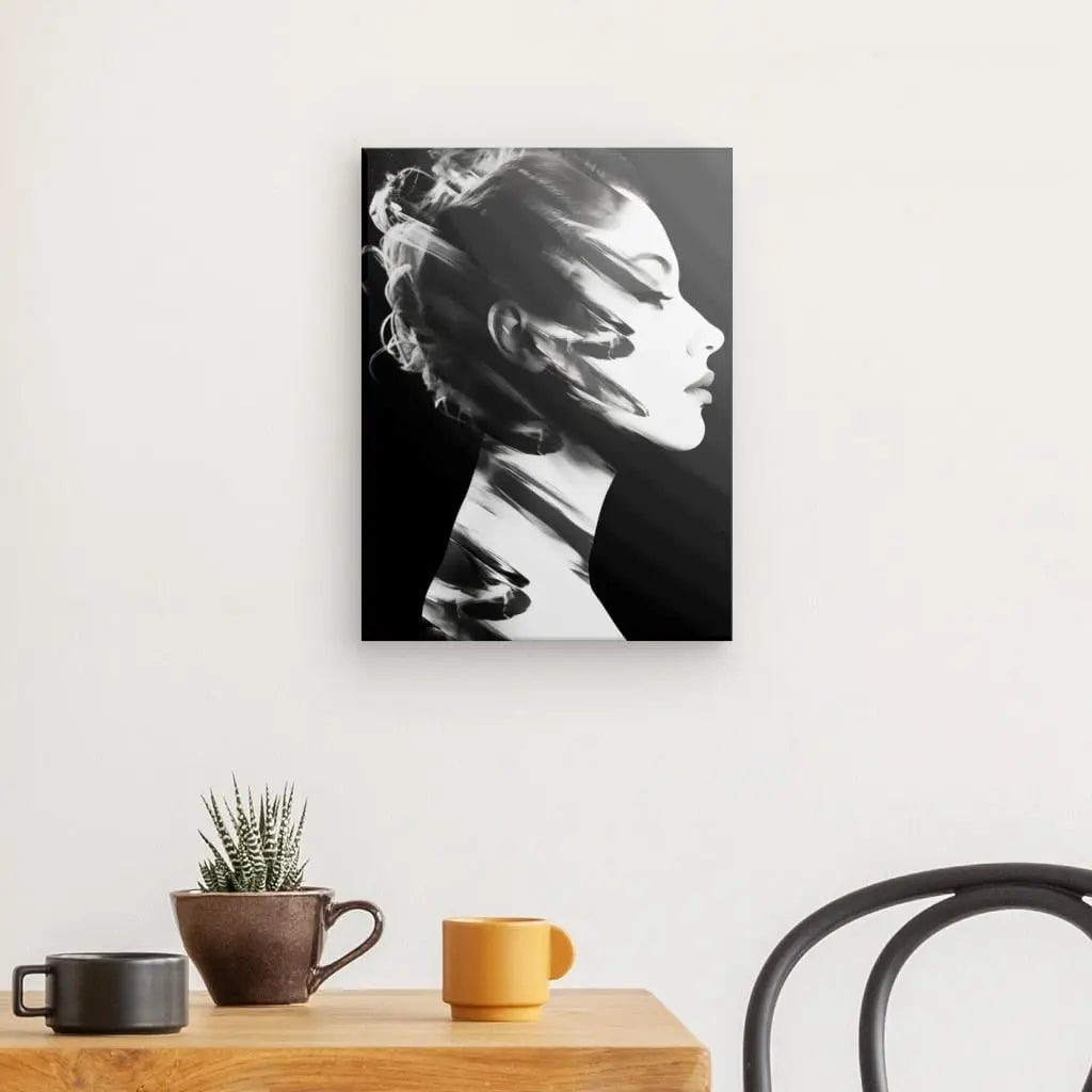 Un Tableau Profil Femme Moderne Noir et Blanc représentant une femme moderne aux cheveux flottants est accroché sur un mur uni au-dessus d'une table qui présente une petite plante en pot, une tasse marron et une tasse jaune. À droite, la vue partielle d’une chaise noire renforce l’ambiance art contemporaine de l’espace.