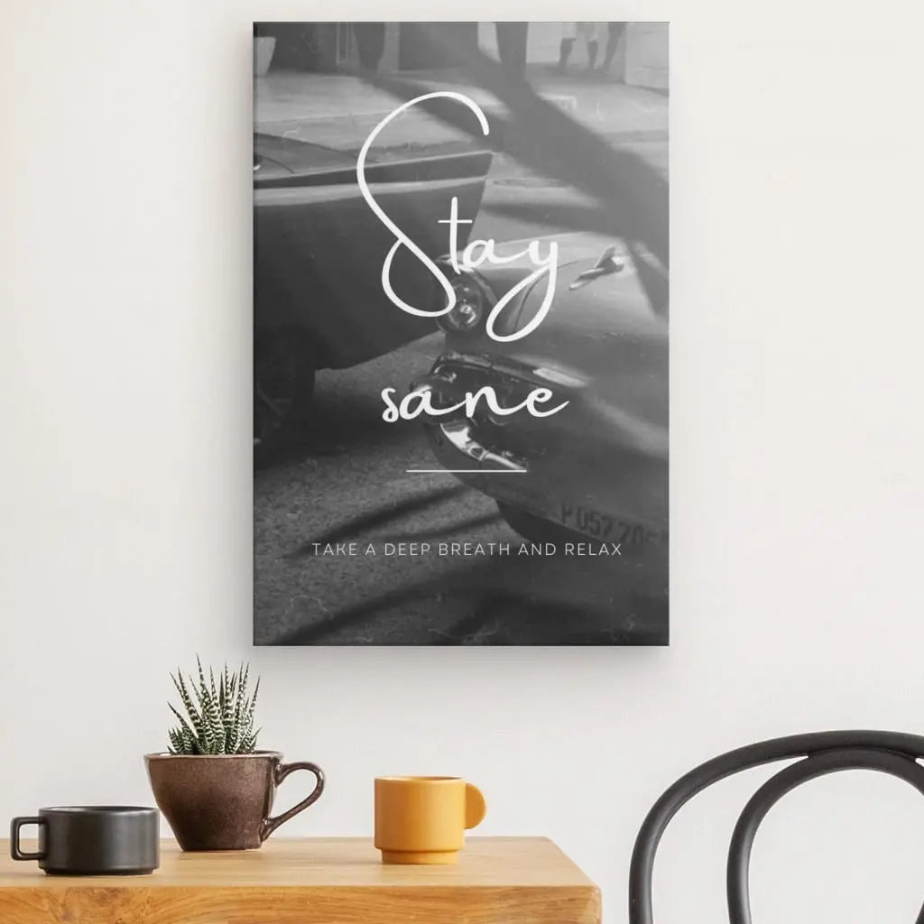 Une affiche Tableau Citation "Stay Sane" Noir et Blanc avec les textes "Stay sain d'esprit" et "Respirez profondément et détendez-vous" est accrochée à un mur au-dessus d'une table en bois ornée de plantes en pot, de deux tasses à café et d'un élégant tableau de citation.