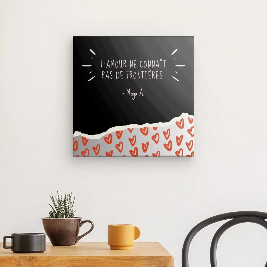 Un mur présentant le Tableau Citation "L'amour n'a pas de frontières" en français, placé au-dessus d'une table avec une plante en pot, une tasse et une tasse à côté d'une chaise noir et blanc.