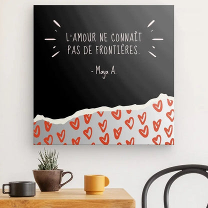 Un Tableau Citation noir et blanc « L'amour n'a pas de frontières » affiche la phrase « L'amour ne connaît pas de frontières. - Maya A. » en français, au-dessus d'un motif de coeurs rouges. Il est monté au-dessus d'une table en bois avec une petite plante en pot et une tasse.