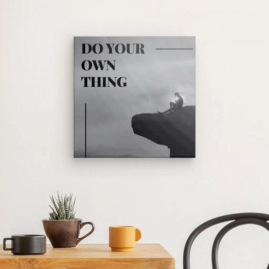 Un Tableau Citation Falaise Noir et Blanc avec le texte « DO YOUR OWN THING » présente une personne assise au bord d'une falaise, imprimé sur une toile tendue à la main à l'aide d'encres latex HP pour des couleurs éclatantes. L'œuvre est suspendue au-dessus d'une table en bois ornée d'un cactus en pot et de deux tasses. Une chaise est partiellement visible.