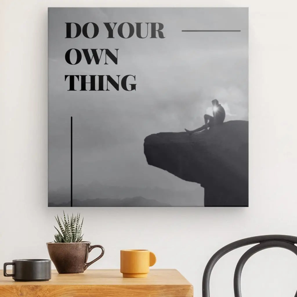 Image en noir et blanc d'une personne assise au bord d'une falaise, avec le texte « DO YOUR OWN THING » en caractères gras. La scène, imprimée avec des encres HP latex, est affichée sous la forme Tableau Citation Falaise Noir et Blanc au-dessus d'une table avec des tasses à café et une petite plante.