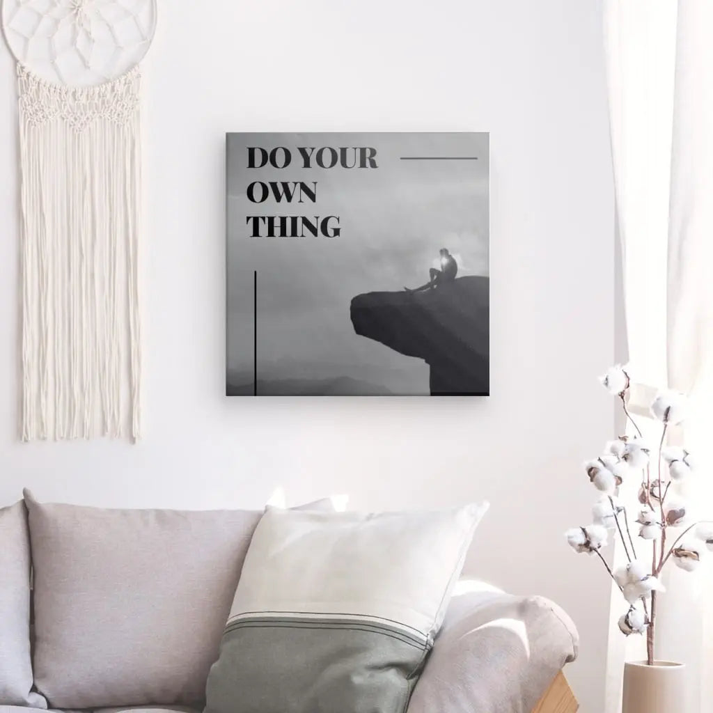 Une image encadrée avec le texte « DO YOUR OWN THING » est accrochée sur un mur blanc au-dessus d'un canapé gris clair avec des oreillers blancs et gris. Une tenture murale et un vase aux branches décoratives sont également visibles. Le Tableau Citation Falaise Noir et Blanc, créé à l'aide d'encres latex HP, dégage des couleurs vibrantes qui rehaussent l'élégance de la pièce.