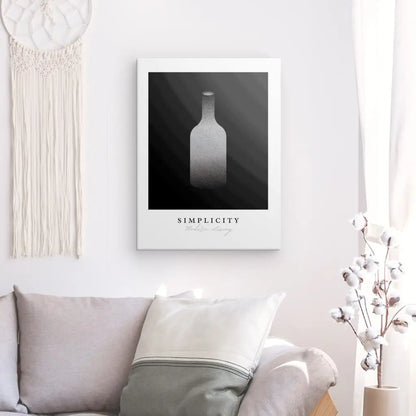 Un espace de vie minimaliste présente une impression d'art encadrée intitulée Tableau Bouteille Design "Simplicity" Noir et Blanc accrochée à un mur blanc, à côté d'un attrape-rêves. En dessous se trouve un canapé gris clair orné d'oreillers, respirant la Décoration Sophistiquée.