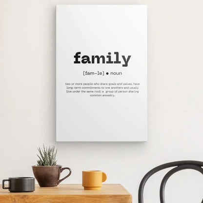 Art mural minimaliste, un Tableau Définition "Famille" Noir et Blanc avec la définition de "famille" en typographie élégante, affiché sur un fond blanc. Il est suspendu au-dessus d'une table en bois ornée de plantes et de deux mugs, à côté d'une élégante chaise noire – parfait pour ceux qui aiment la décoration noire et blanche.