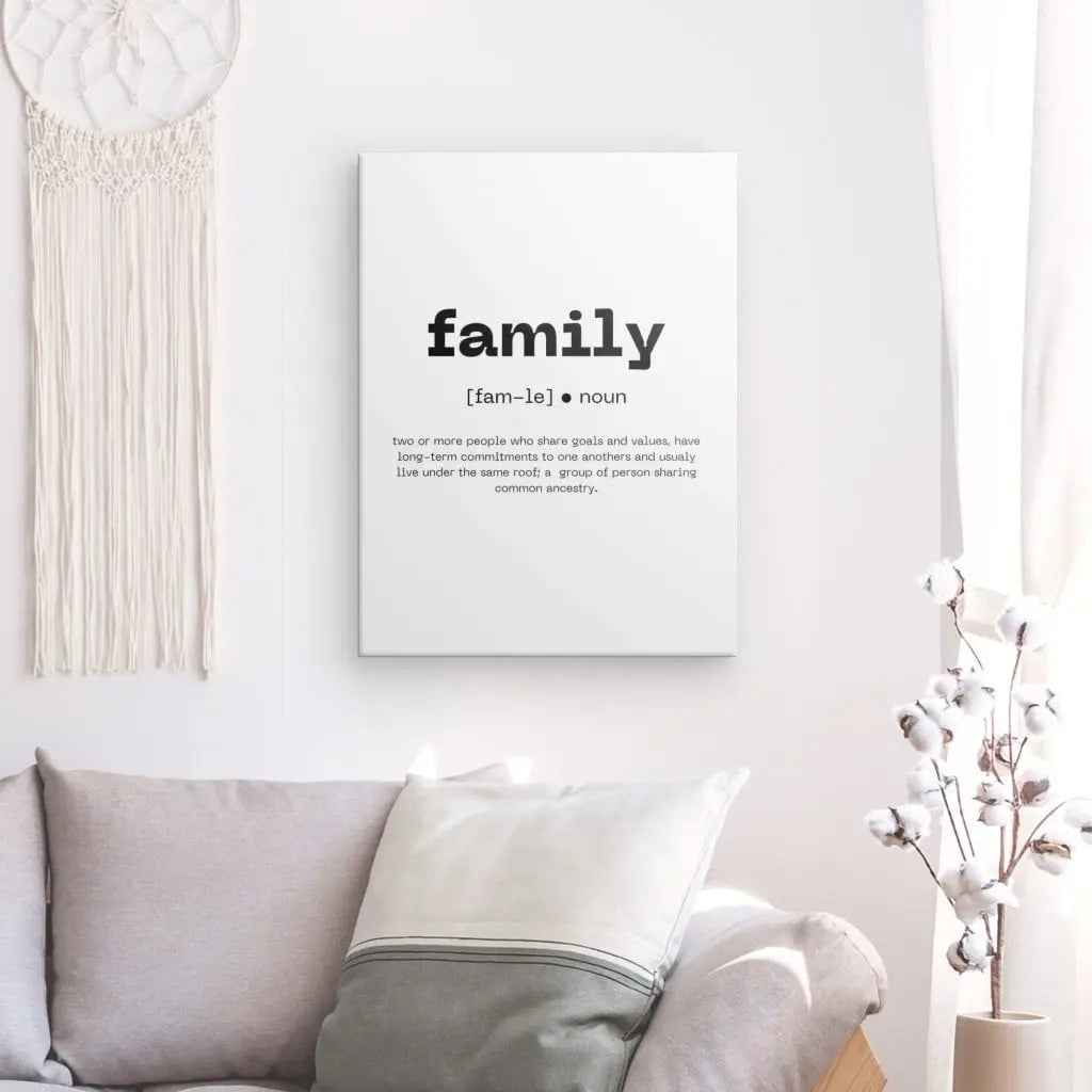 Chambre minimaliste avec un Tableau Définition "Famille" Noir et Blanc à côté d'une impression encadrée définissant le mot "famille" en typographie élégante. Un canapé de couleur claire avec des coussins est placé en dessous, complété par une plante à fleurs blanches sur la droite. La décoration noire et blanche ajoute à l'ambiance sereine.