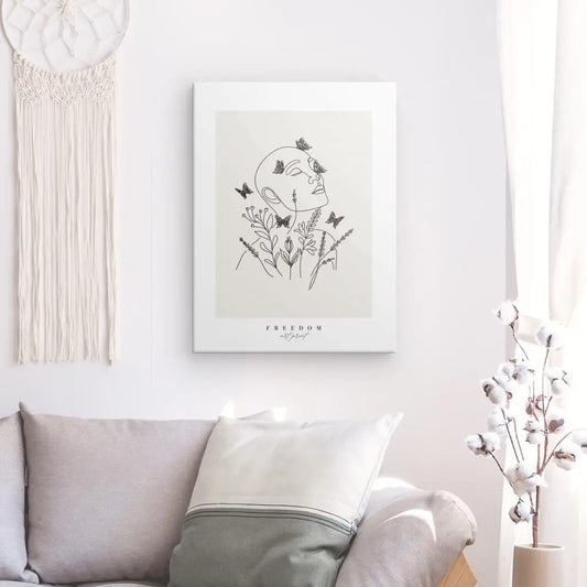 Une illustration minimaliste d'un visage entouré de fleurs et de papillons, intitulée "Tableau 'Freedom' Beige Noir et Blanc", est accrochée sur un mur blanc au-dessus d'un canapé beige avec des oreillers. Un attrape-rêves et une plante sont de chaque côté, ajoutant à la décoration harmonieuse.