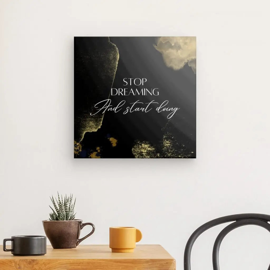 Un mur avec une citation encadrée « Arrêtez de rêver et commencez à faire » est suspendu au-dessus d'une table en bois contenant une plante en pot, une tasse orange et une tasse marron. Le Tableau "Stop Dreaming" Design Noir Blanc et Doré ajoute de l'élégance à côté de la configuration. Une chaise noire est placée à côté de la table.